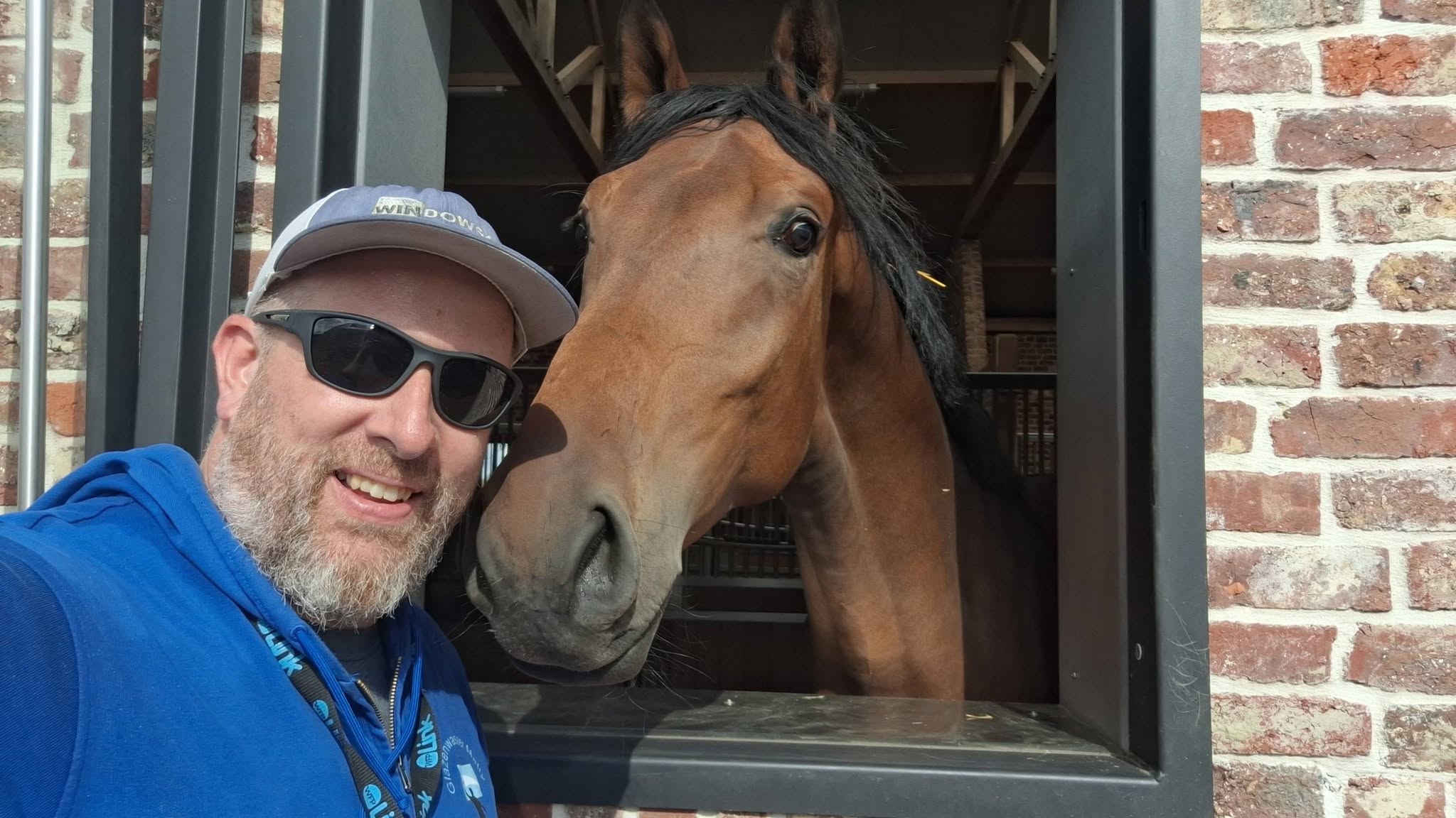 Matty met een pet en zonnebril naast een paard dat uit een stalraam kijkt