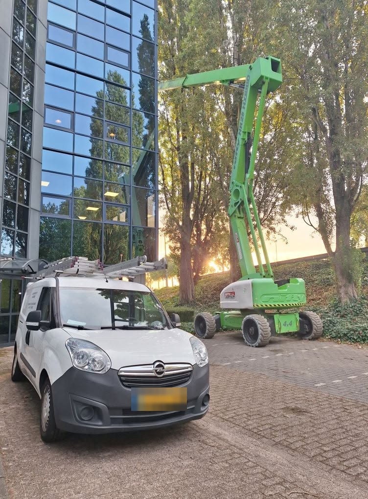 Matty's witte bestelbus naast een groene hoogwerker bij een gebouw met glazen gevel