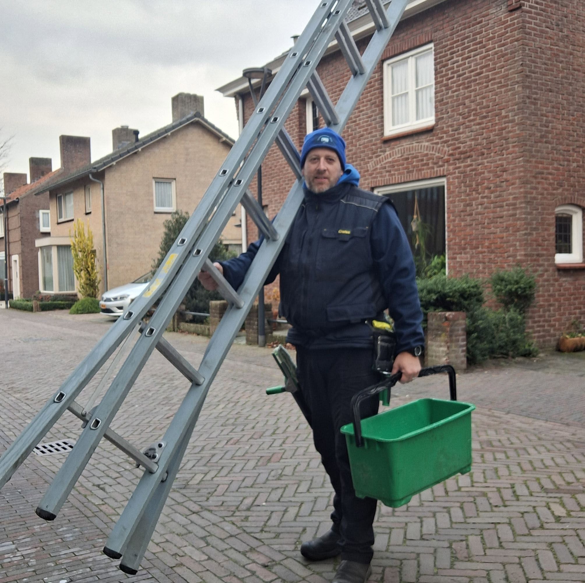 Matty met een ladder op zijn schouder en een groene emmer in de hand, in een straat in de buurt