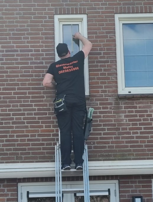Matty op een ladder bij een raam op de eerste verdieping van een bakstenen woning