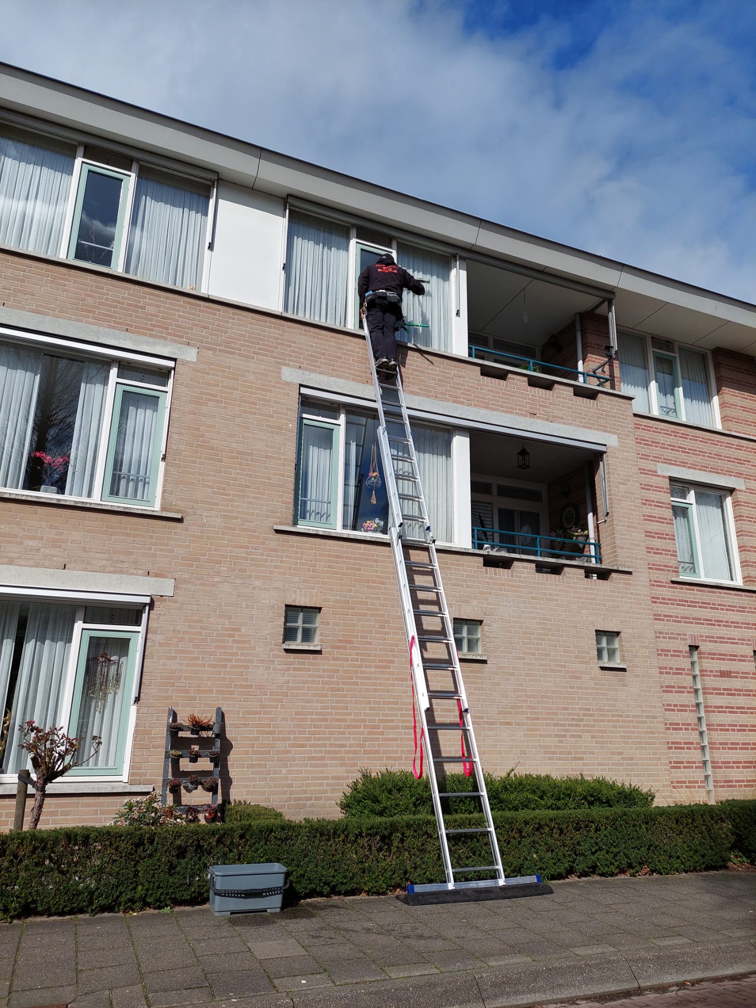 Matty hoog op een lange ladder tegen een appartementencomplex, bij een raam op de tweede verdieping